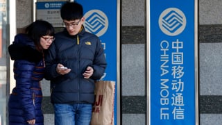 Eine Chinesin und ein Chinese stehen vor zwei China Mobile-Plakaten und schauen auf ein iPhone.