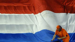 Eine orange gekleidete Frau vor einer niederländischen Flagge.