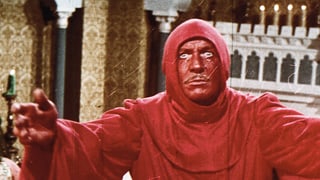 Filmstill: Ein Mann mit rot angemaltem Gesicht in einem roten Kapuzenkleid 