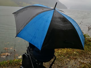 Ein  Mensch mit  einem Schirm, sitzt am Ufer eines Sees, es regnet in strömen. 