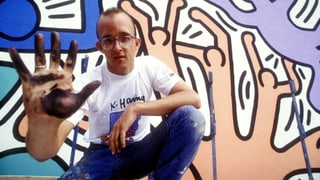 Keith Haring vor einer Wand, die er bemalt hat, die mit Farbe verschmierte Hand in die Kamera haltend.