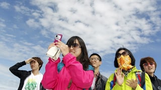 Gruppe von fünf asiatischen Touristen vor blauem Himmel mit ein paar Wolken, eine Frau macht ein Foto.