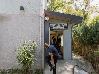 Ein Eingangsbereich eines Hauses mit grauen Wänden. Drinnen sind einige Männer zu sehen, vor der Türe zieht sich ein Mann die Schuhe an. Ein Eingangsbereich eines Hauses mit grauen Wänden. Drinnen sind einige Männer zu sehen, vor der Türe zieht sich ein Mann die Schuhe an.