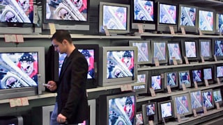 Ein Mann schaut vor einer Wand von TV-Bildschirmen auf sein Handy.