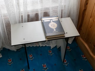 Auf einem kleinen Tisch vor einem Vorhang liegt ein Koran, ein grosses, schwarzes Buch mit goldenen Verzierungen.