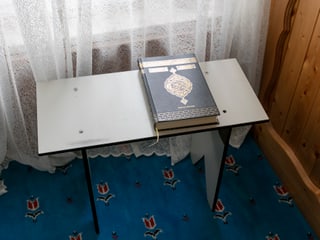 Auf einem kleinen Tisch vor einem Vorhang liegt ein Koran, ein grosses, schwarzes Buch mit goldenen Verzierungen. Auf einem kleinen Tisch vor einem Vorhang liegt ein Koran, ein grosses, schwarzes Buch mit goldenen Verzierungen.