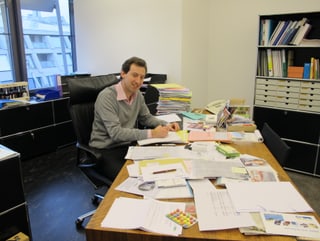 Ein Mann in grauem Pullover und rosa Hemd sitzt an einem Tisch mit vielen Dokumenten.