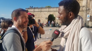 Ein Mann mit heller Hautfarbe streckt einem anderen Mann mit dünklerer Hautfarbe ein Mikrophon entgegen. Hinter ihnen ist eine Kamera zu erkennen, die auf den Interviewten gerichtet ist. Ein Mann mit heller Hautfarbe streckt einem anderen Mann mit dünklerer Hautfarbe ein Mikrophon entgegen. Hinter ihnen ist eine Kamera zu erkennen, die auf den Interviewten gerichtet ist.