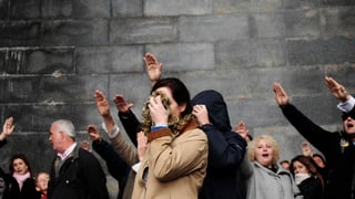 Mehrere Personen mit Faschistengruss stehen vor einer grauen Wand, eine Frau verdeckt ihr Gesicht.