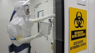 Symbolbild: Biogefahren-Symbol und eine Person hinter einer Schleuse in Schutzkleidung. Symbolbild: Biogefahren-Symbol und eine Person hinter einer Schleuse in Schutzkleidung.
