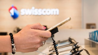 Eine Hand hält ein Smartphone, im Hintergrund das Swisscom-Logo.