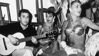 Gitarrist Joao Gilberto sitzt neben zwei Tänzerinnen, die mit Federn geschmückt sind.