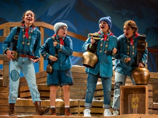 Vier Kinder in traditioneller Schweizer Kleidung stehen auf Holzkisten und singen. Alle tragen eine Glocke um den Arm. Sie spielen Kinder aus dem Kinderbuchklassiker Schellen-Ursli Vier Kinder in traditioneller Schweizer Kleidung stehen auf Holzkisten und singen. Alle tragen eine Glocke um den Arm. Sie spielen Kinder aus dem Kinderbuchklassiker Schellen-Ursli