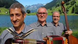 Die drei Musiker mit ihren Instrumenten vor einem Bergsee.