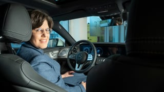Eine Frau mit blauem Anzug sitzt im Auto und hält mit der linken Hand das Lenkrad. Sie grinst über die Schulter nach hinten in die Kamera. Eine Frau mit blauem Anzug sitzt im Auto und hält mit der linken Hand das Lenkrad. Sie grinst über die Schulter nach hinten in die Kamera.