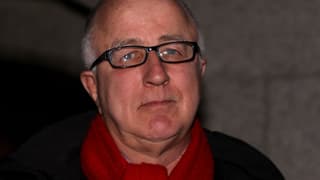 Porträtaufnahme von MacShane mit rotem Schal und schwarzer Brille.
