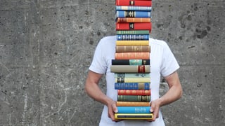 Ein Mann im weissen T-Shirt trägt einen so grossen Bücherstapel, dass man seinen Kopf nicht mehr sieht.