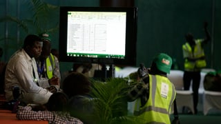 Wahlhelfer befinden sich in einem Gebäude. Auf einem grossen Bildschirm werden die Wahlergebnisse aktualisiert.