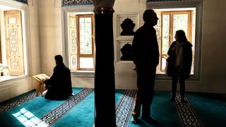 In einem kleinen Gebetsraum: Zwei Besucher sehen sich am Tag der offenen Türe die Berliner Sehitlik Moschee an, während ein Muslime darin betet.