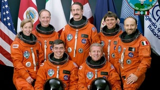 Sechs Astronauten und eine Astronautin tragen auf dem Gruppenbild orange Weltraumanzüge.