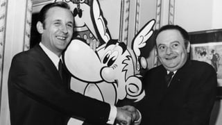 Albert Uderzo und René Goscinny geben sich die Hand. In ihrer Mitte steht eine Asterix-Karton-Figur.