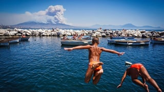 Rauchender Vesuv in der Ferne, im Vordergrund springen zwei Badende Jungs ins Meer.