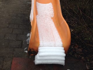 Eine orange Kinderrutsche. Wie zusammengefaltet türmt sich am Fusse der Rutsche der Schnee.