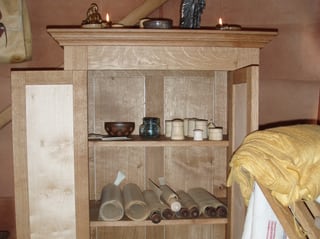 Doktorschrank mit Schriftrollen, mehreren weissen Gefässen aus Holz und Glasfläschchen. Auf dem Schrank stehen brennende Kerzen.