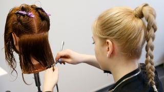 junge Frau mit blondem Zopf von hinten zu sehen, wie sie einer Puppe die Haare schneidet