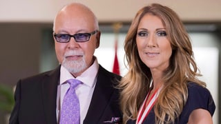 René Angélil und Celine Dion Halbnahe lachen in Kamera. Er mit Brille, Anzug und violetter Krawatte. Sie im schwarzen Oberteil und Medaille um Hals.