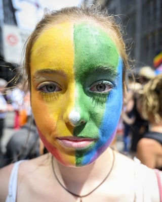 Eine junge Frau mit in Regenbogenfarben angemaltem Gesicht blickt in die Kamera.
