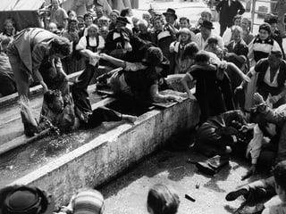 Tumult auf dem Dorfplatz. Die Dorfbewohner stehen um einen Brunnen, in welchem sich eine Handvoll Männer raufen.