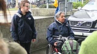 Ein junger Mann mit einer Umhängetasche auf der linken Bildseite, eine ältere Frau im Rollstuhl auf der rechten Bildseite vor einem Auto.