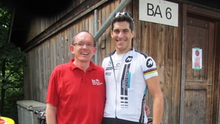 Radrennprofi Franco Marvulli trägt ein weisses Trikot mit Werbung darauf, sowie eine Sonnenbrille, die er in die Haare gesteckt hat. André Keller trägt ein rotes Polo-Shirt und eine normale Brille. Zusammen posieren sie vor einer alten Scheune. Radrennprofi Franco Marvulli trägt ein weisses Trikot mit Werbung darauf, sowie eine Sonnenbrille, die er in die Haare gesteckt hat. André Keller trägt ein rotes Polo-Shirt und eine normale Brille. Zusammen posieren sie vor einer alten Scheune.