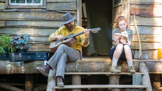 Filmstill: Ein Mann spielt Gitarre, ein Mädchen liest ein Buch.