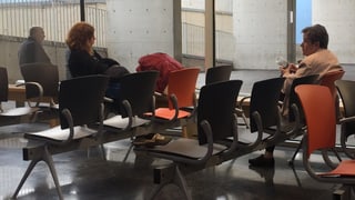 Das Wartezimmer des Hospital del Mar in Barcelona. Man sieht zwei Frauen und einen älteren Herrn, die auf orangenen Stühlen warten. Ansonsten wirkt der Raum leer und kalt. 