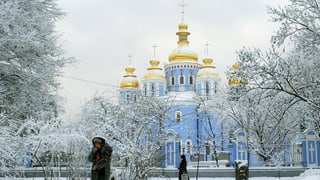 Ältere Frau stapft durch den Schnee vor einer Kathedrale mit goldenen Kuppeln.