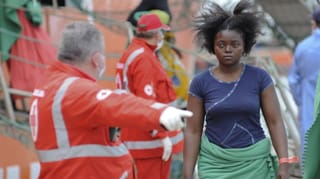 Ein Mitarbeiter des IKRK in rotem Anzug und mit Mundschutz weist einer jungen Afrikanerin einen Platz zu.