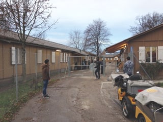 Das Bild zeigt die Schule, die in einer Art Baracken sind. Sie sind eingezäunt mit einem hohen Draht und einem grossen Tor. Die Bäume haben keine Blätter mehr. Es stehen einige junge Menschen vor der Türe. 