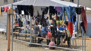 Flüchtlinge, die ihre Wäsche aufgehängt haben und im Freien an Festbänken sitzen.