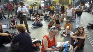 Passanten sitzen auf dem Perron am Boden und vertreiben sich die Zeit mit Lesen.