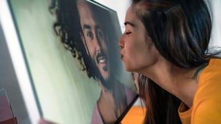 Eine Frau küsst einen Bildschirm, auf dem ein Mann zu erkennen ist.