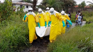 Helfer in Schutzkleidung tragen in einem weissen Tuch ein totes Ebola-Opfer aus einem Dorf.
