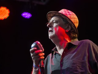 Mann mit Mikrofon in der Hand und Hut auf dem Kopf vor einer schwarzen Wand