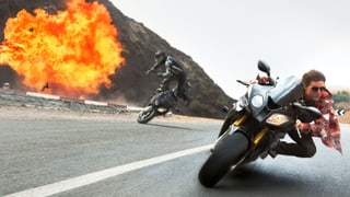 Tom Cruise auf dem Motorrad in einer Kurve und hinten sieht man eine Explosion.