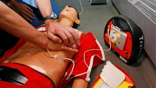 Ein Mann verabreicht einer Puppe eine Herzmassage; daneben steht ein Defibrillator.