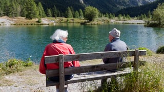 Eine älteres Paar sitzt auf einer Bank an einem Bergsee.
