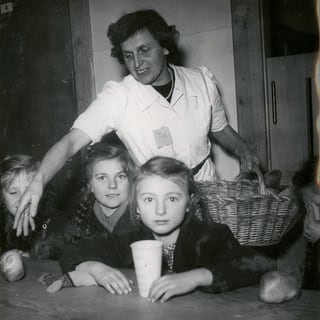 Eine Frau steht hinter Kindern, die an einem Tisch sitzen.