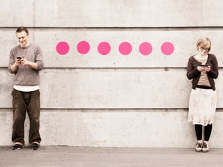 Ein Mann und eine Frau stehen an einer Wand und schauen jeweils auf einen Bildschirm. Eine Linie aus rosa Punkten verbindet ihre Körper. 