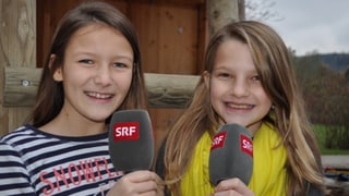 Annika und Sophie stehen im Freien und halten je ein Mikrofon in den Händen. Annika und Sophie stehen im Freien und halten je ein Mikrofon in den Händen.